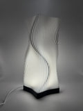 Moderne weiße Lampe mit 3D-gedruckter, wellenförmiger Lamellenstruktur, die ein indirektes Licht  wirft.
